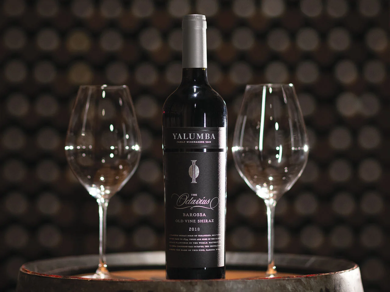The Octavius Shiraz bottle with two empty wine glasses against wooden barrel background