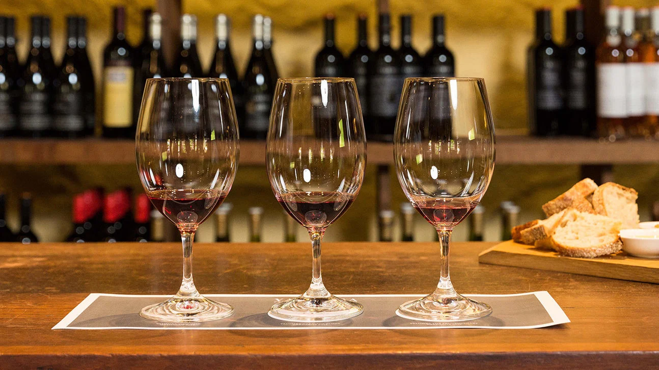 Three glasses of red wine sitting on a bar in front of a large wine rack in the tasting room